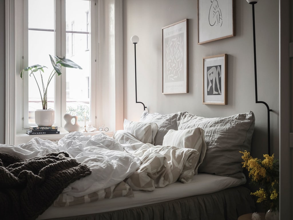 A bedroom with grey walls, a pholc wall lamp, gallery wall, linnen bedding, bedskirt, plant in the window