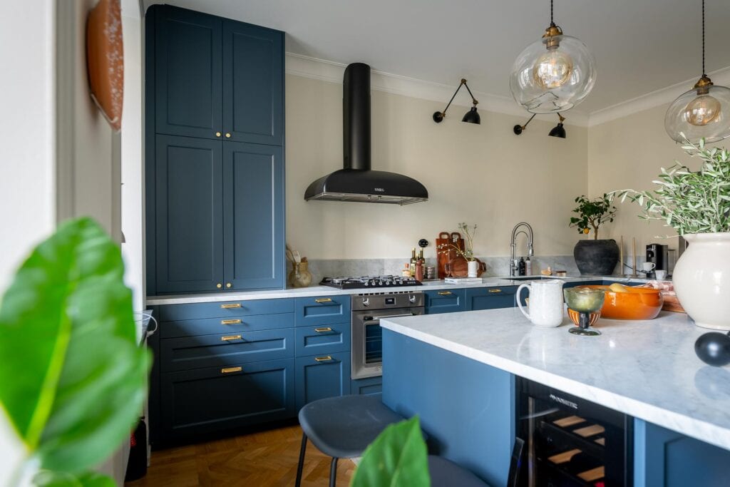 A blue shaker kitchen with gold hardware, a kitchen island, white marble countertops, stainless steel appliances, black wall lamps