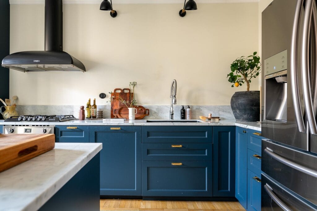 A blue shaker kitchen with gold hardware, a kitchen island, white marble countertops, stainless steel appliances, black wall lamps