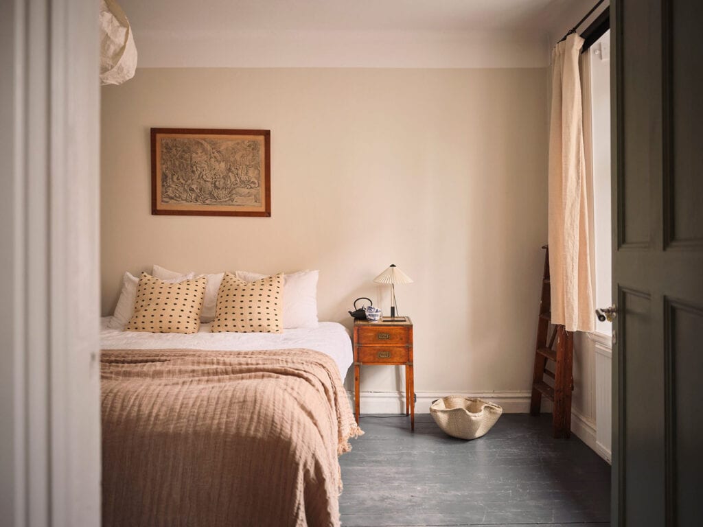 A bedroom with beige walls, grey flooring, a mixture of beige and white textiles and two different vintage bedside tables