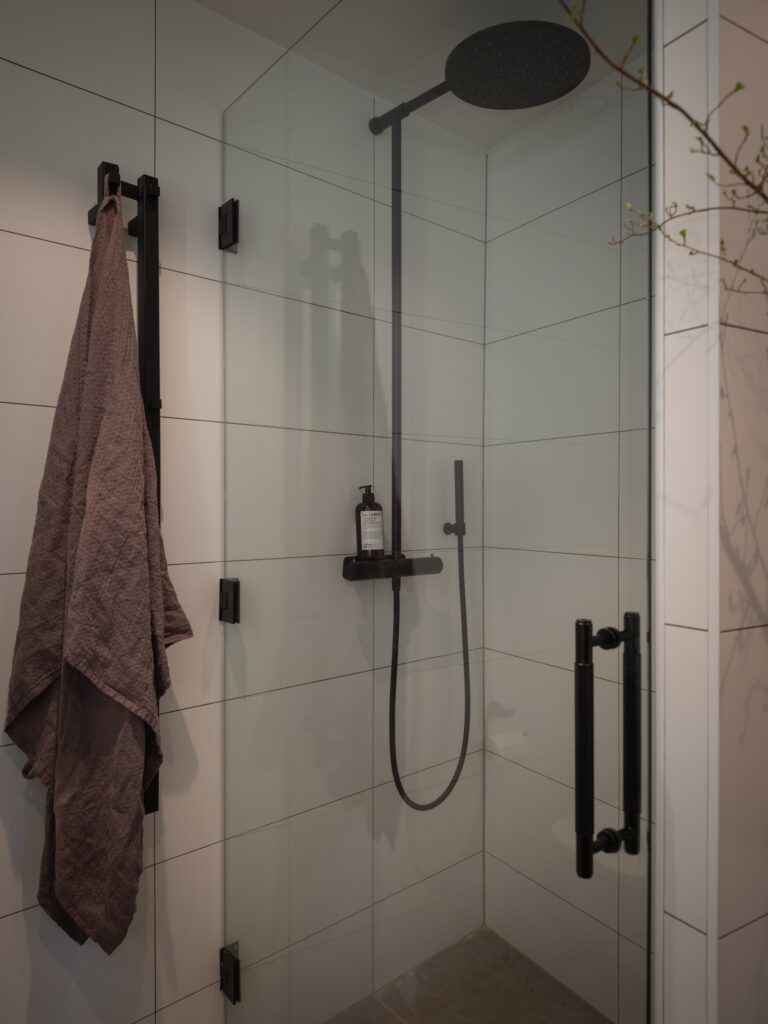 A modern bathroom with white wall tiles, a glass shower door, and black fixtures