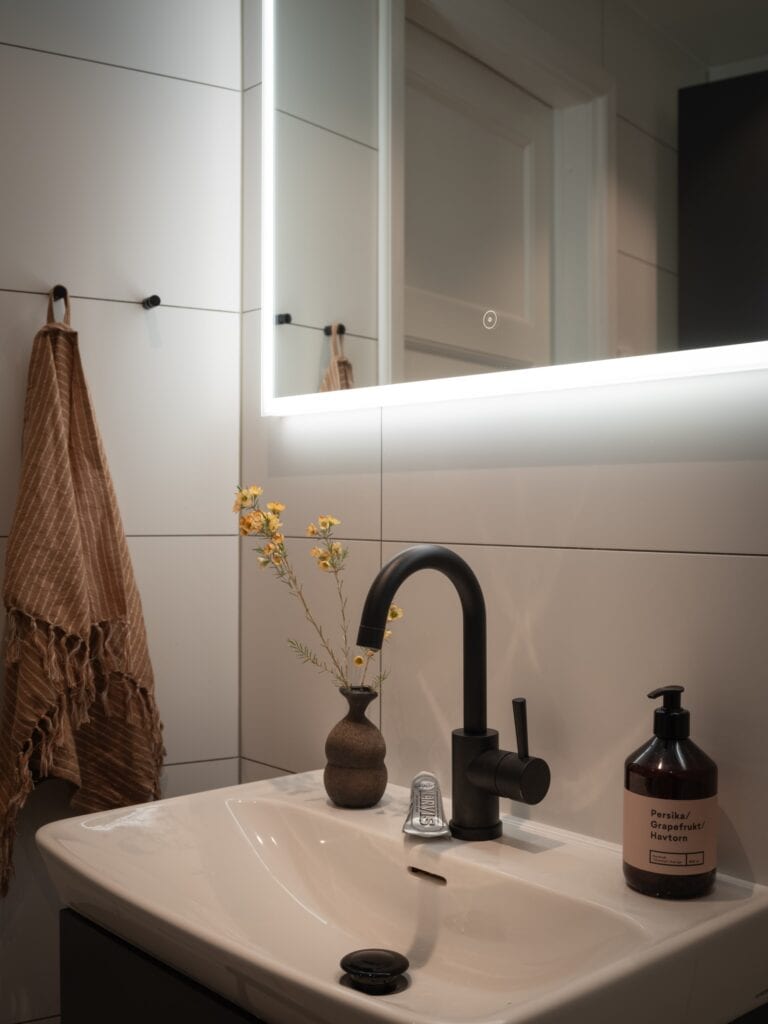 A modern bathroom with white wall tiles, an LED mirror and black fixtures