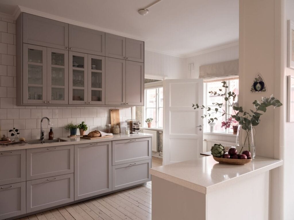 A light grey kitchen with a white countertop, glass upper cabinets and white tile backsplash