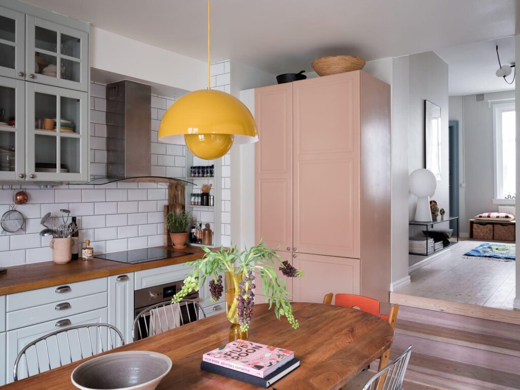 A mint green kitchen with butcher block countertops, a white subway tile backsplash and upper glass cabinets, with a pink cabinet for the fridge