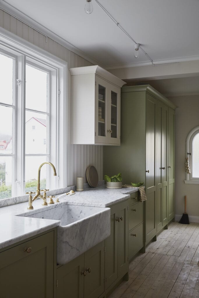A white marble farmhouse sink in an olive green kitchen with brass hardware