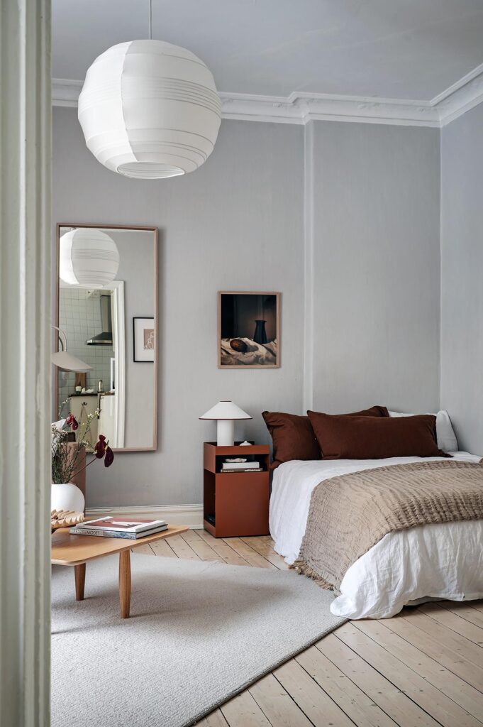 A living and sleeping space with light grey walls and brown and red tones in the textiles, furniture pieces and accessories