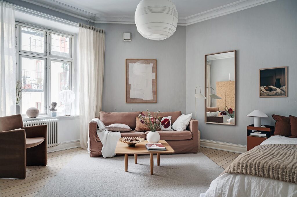 A living and sleeping space with light grey walls and brown and red tones in the textiles, furniture pieces and accessories