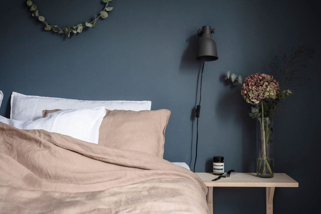 A bedroom with a deep blue wall color, black wall lamps, white and dusty pink bedding