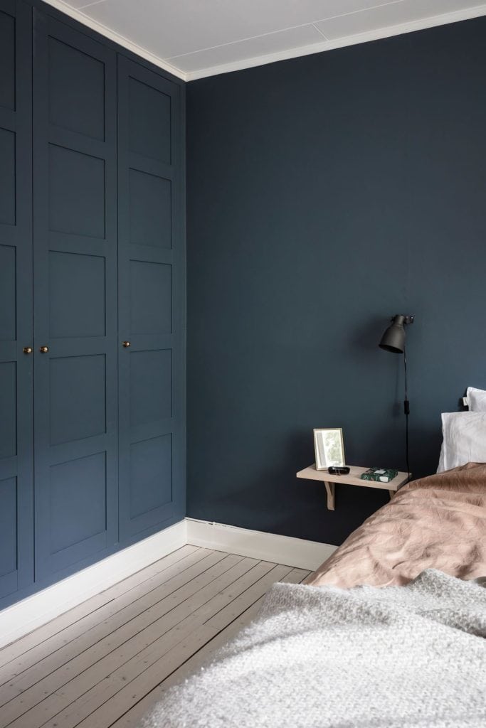 A bedroom with a deep blue wall color, black wall lamps, white and dusty pink bedding, deep blue wardrobe