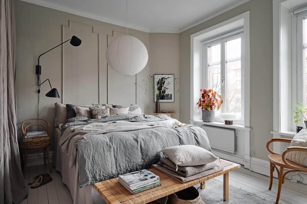 A bedroom with greige walls, beige and gray bedding, wood bench behind the bed, black wall lamp, rice paper shade