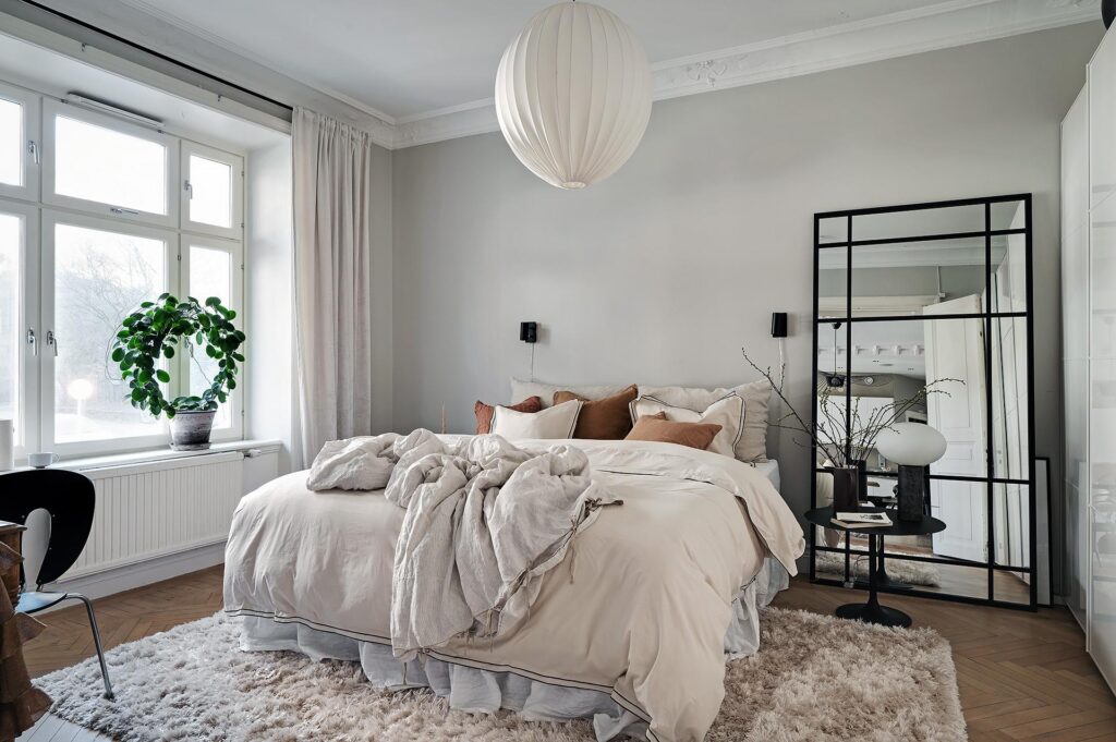 A bedroom with light grey walls, beige bedding, beige fluffy area rug, white pendant, XL mirror