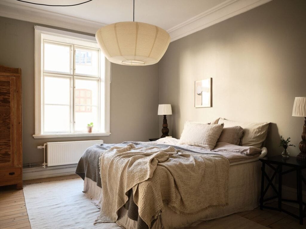 A bedroom with greige walls, beige and gray bedding, rice paper pendant, white area rug