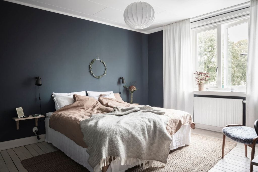 A bedroom with a deep blue wall color, black wall lamps, white and dusty pink bedding, natural rug, white curtains