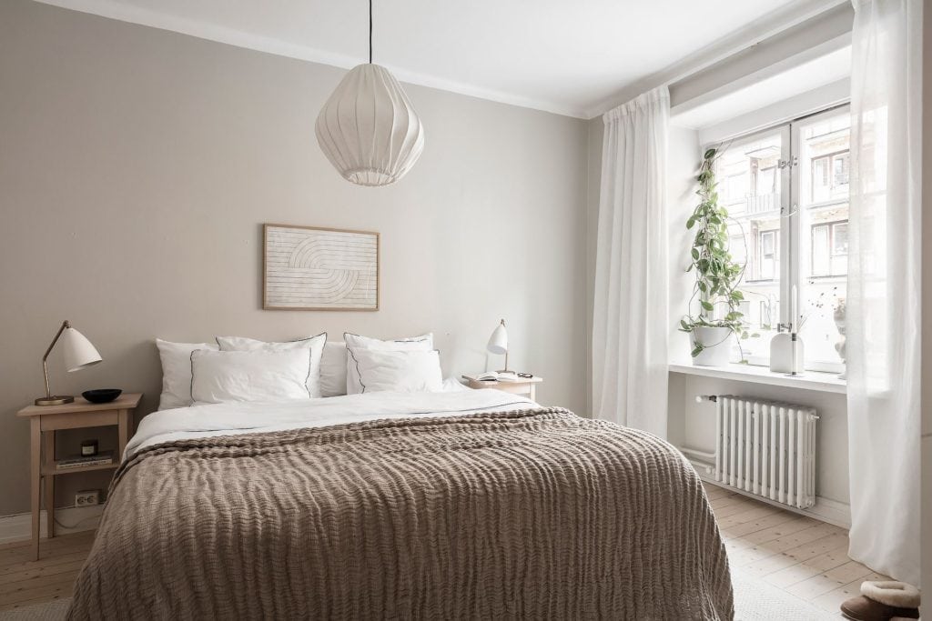 A bedroom with greige walls, beige blanket, white bedding, white curtains, wood bedside tables