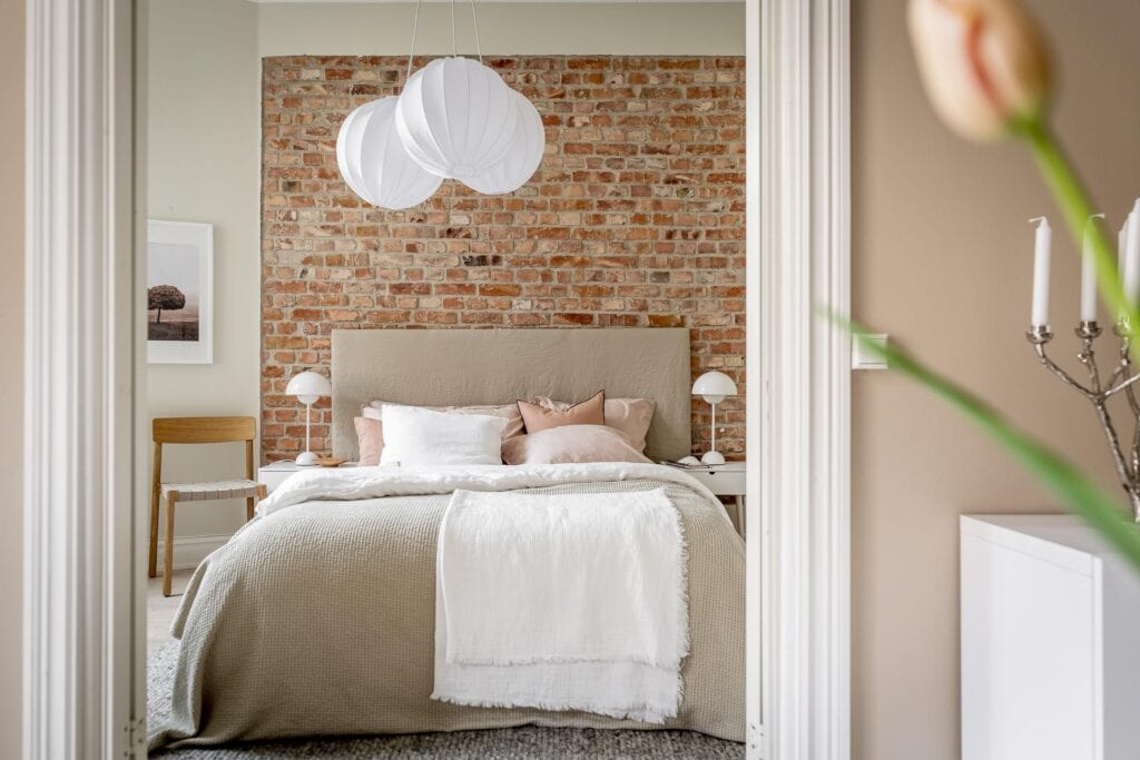 A beige bedroom with an partially exposed brick wall and elegant bedding in beige and pink
