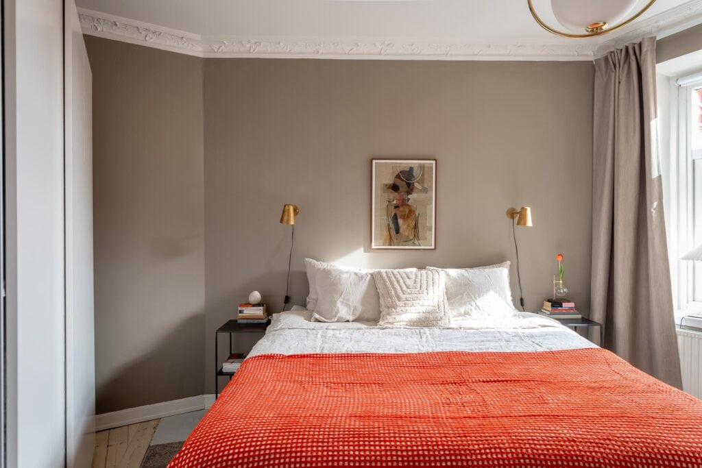 A bedroom with greige walls, orange throw blanket, gold wall lamps, white bedding, beige curtains