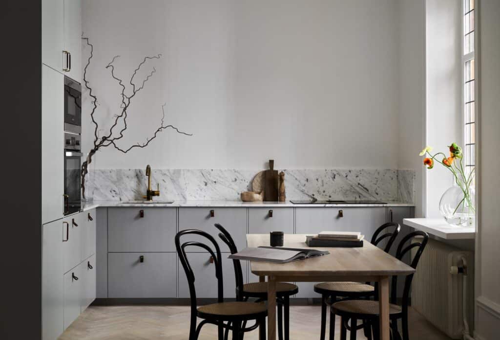 A living kitchen with grey cabinets, a brass faucet and leather handles with a wooden dining table and bentwood chairs
