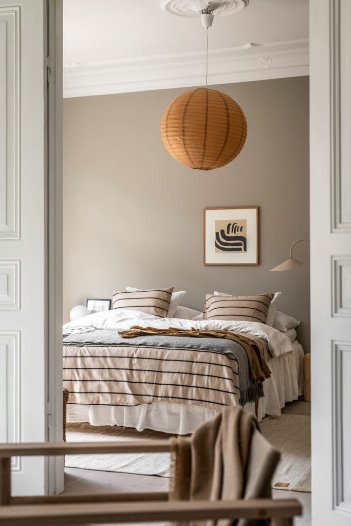 A bedroom with greige walls, a rust-colored rice paper pendant, beige striped bedding, beige Arum floor lamp
