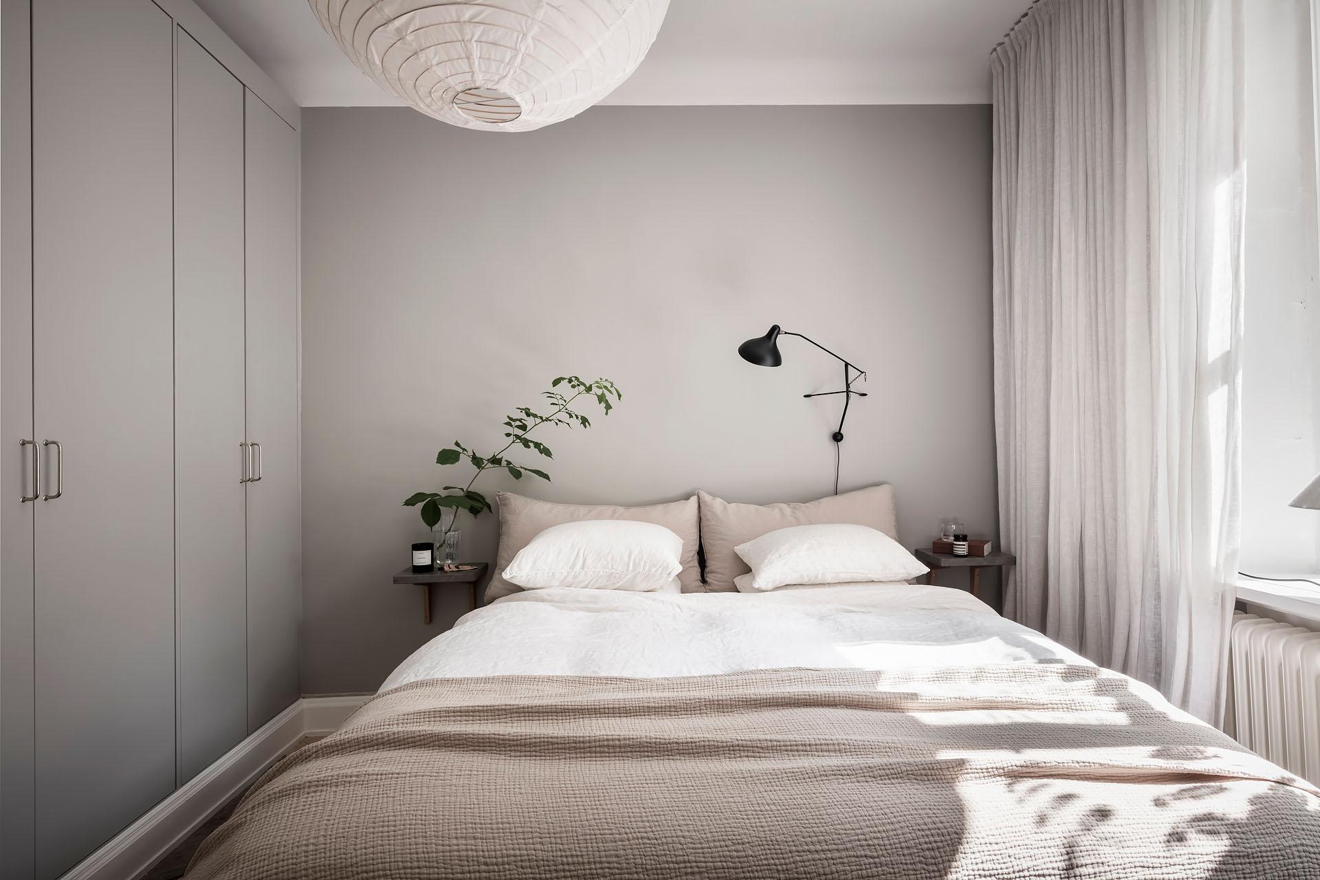 A bedroom with light grey walls, beige bedding, a light grey wardrobe, black wall lamp