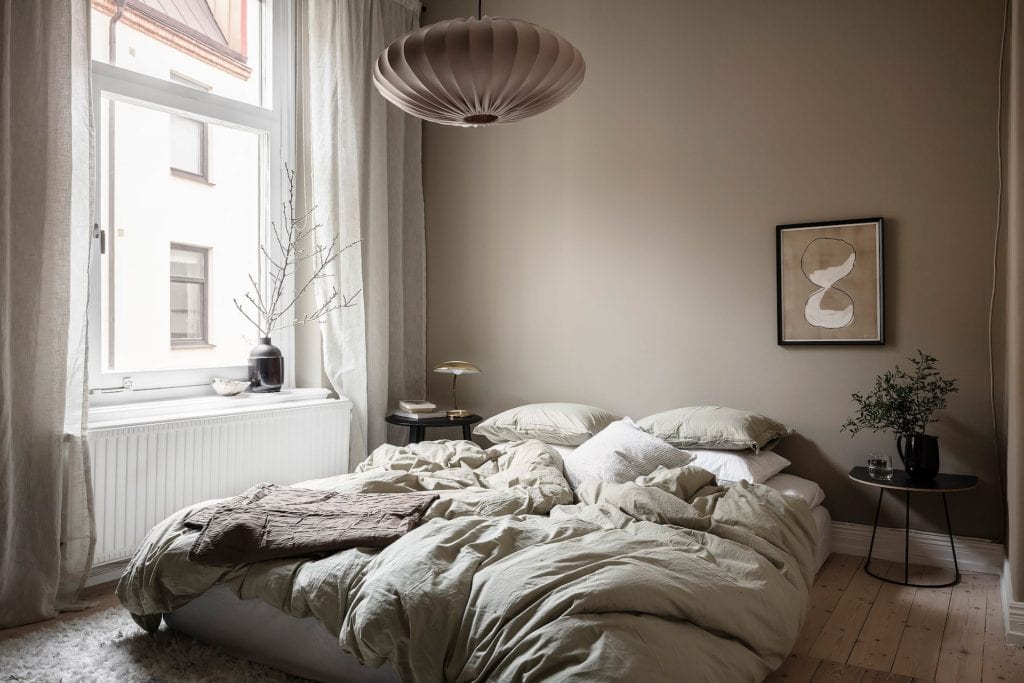 A bedroom with greige walls, sage green bedding, beige artwork, greige linen curtains