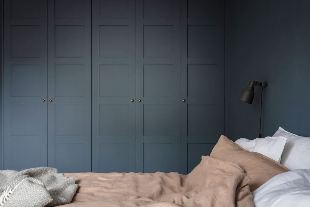 A bedroom with a deep blue wall color, black wall lamps, white and dusty pink bedding, deep blue wardrobe