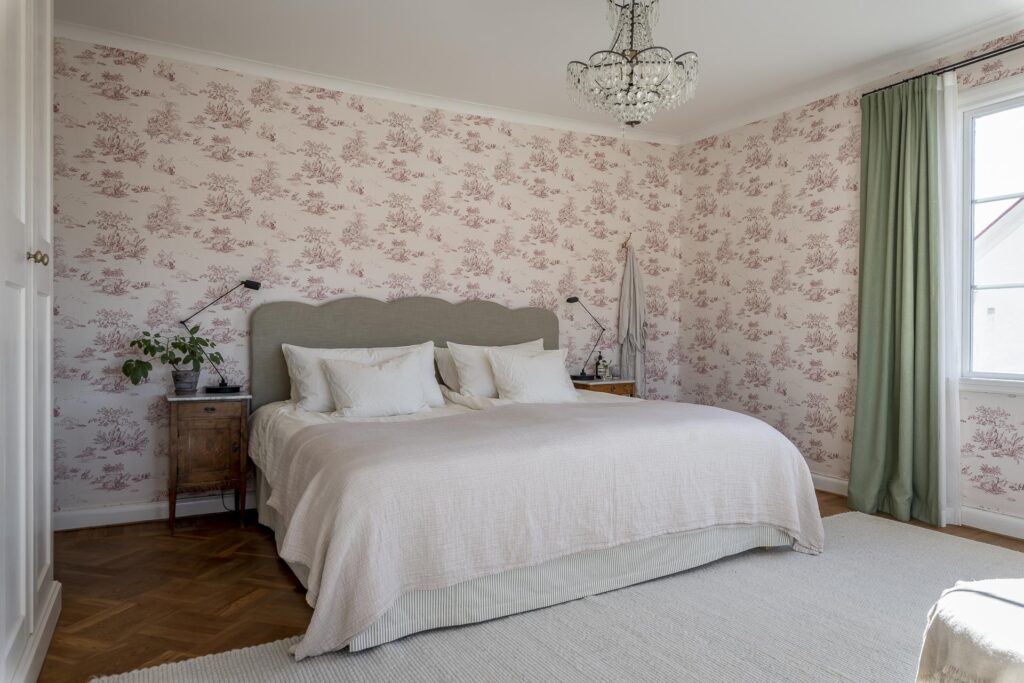 A farmhouse bedroom with a pink bedroom wallpaper, green curtains and a fabric scalloped headboard
