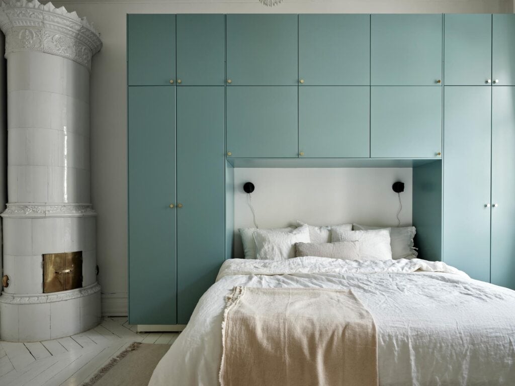 A white bedroom with a blue wardrobe above. the bed, beige curtains, white and beige bedding