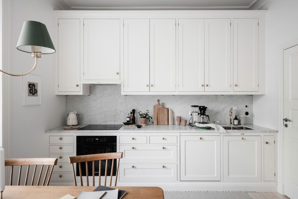 A white shaker kitchen with brass hardware and a white marble countertop and backsplash