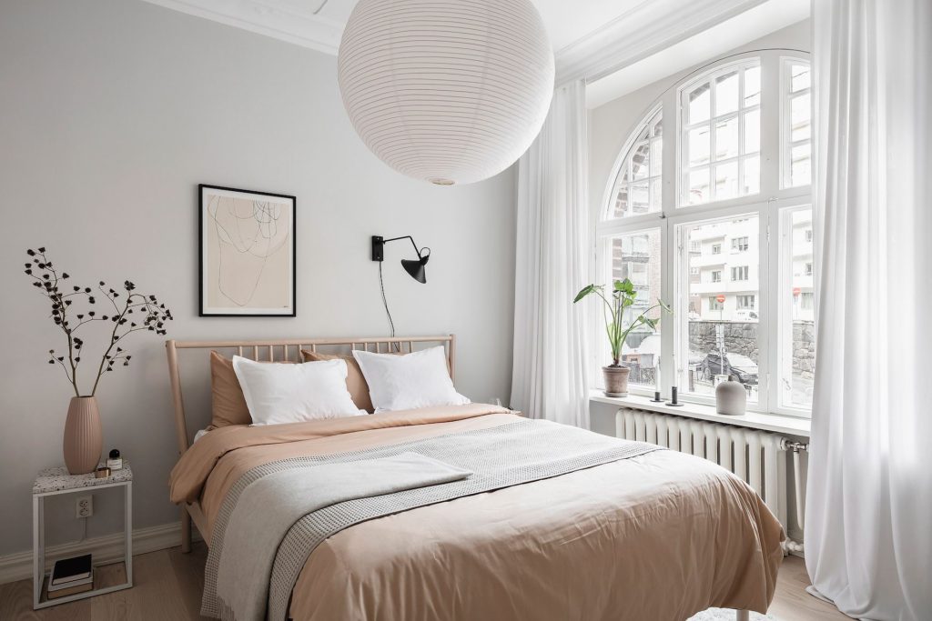 A light grey bedroom with white curtains, nude pink bedding, wood bed frame, rice paper pendant, black wall lamp