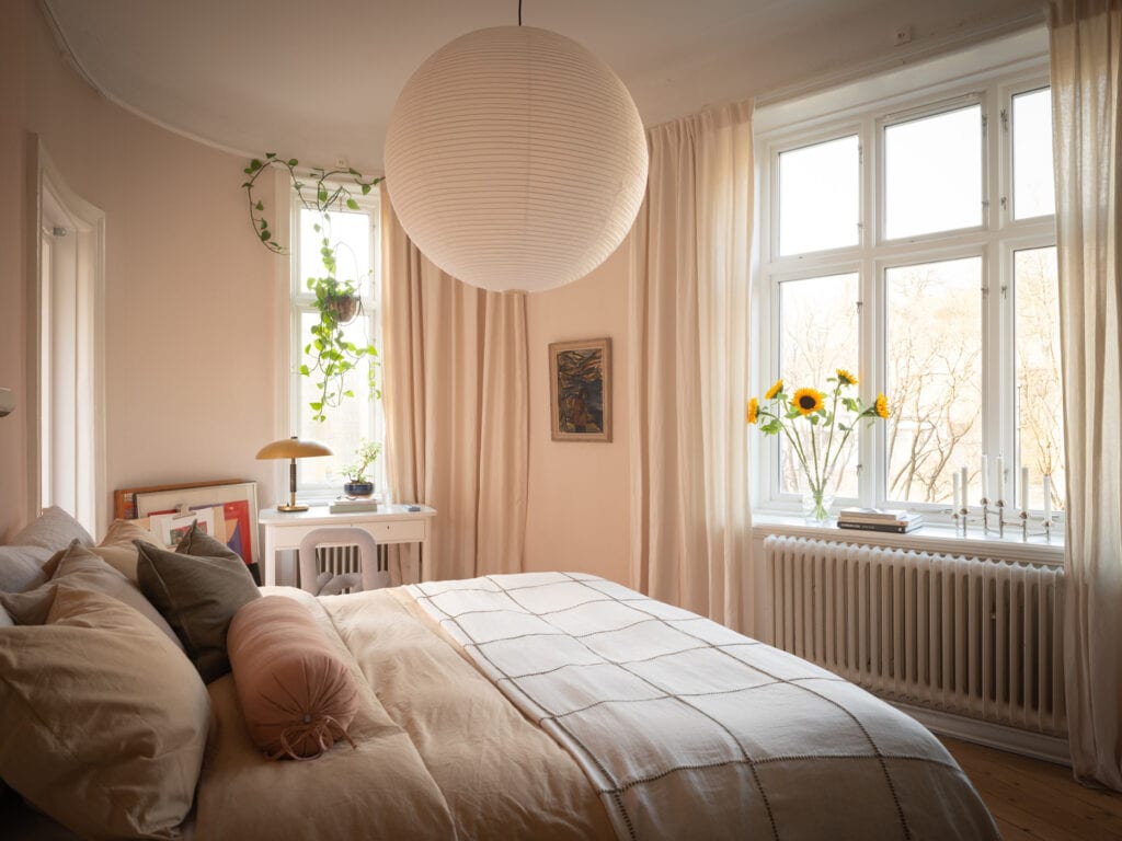 A bedroom with a light pastel pink wall color and light beige curtains