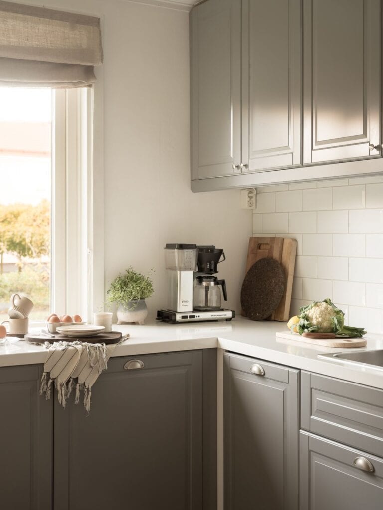 A grey kitchen in a corner layout with chrome hardware, a tick white countertop and a white subway tile backsplash