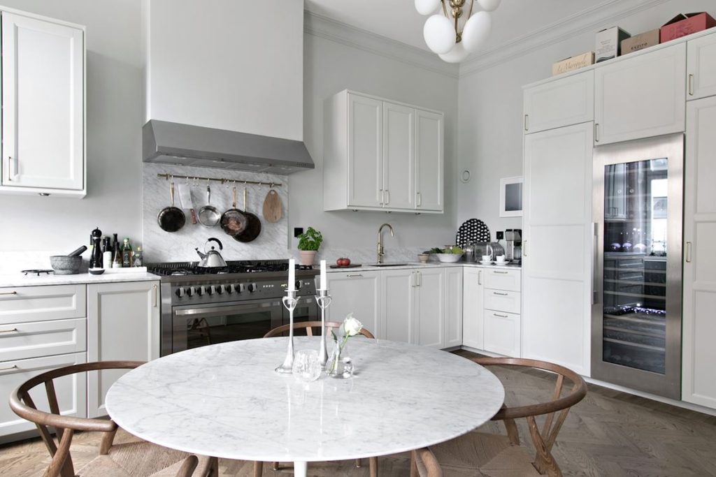 White shaker-style kitchen cabinets, stainless steel stove and hood, upper cabinets, wine cooler, round marble dining table, CH24 wishbone chairs