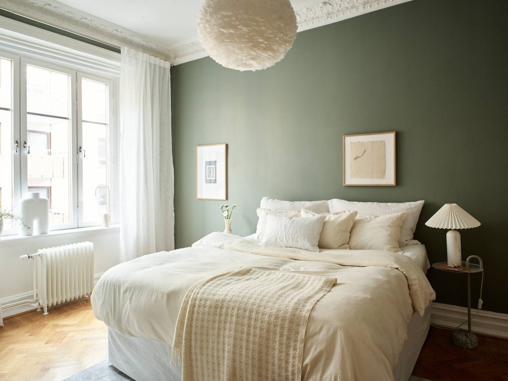 A bedroom with a deep green wall color combined wtih beige bedding, a light grey area rug and a white Umage eos lampshade