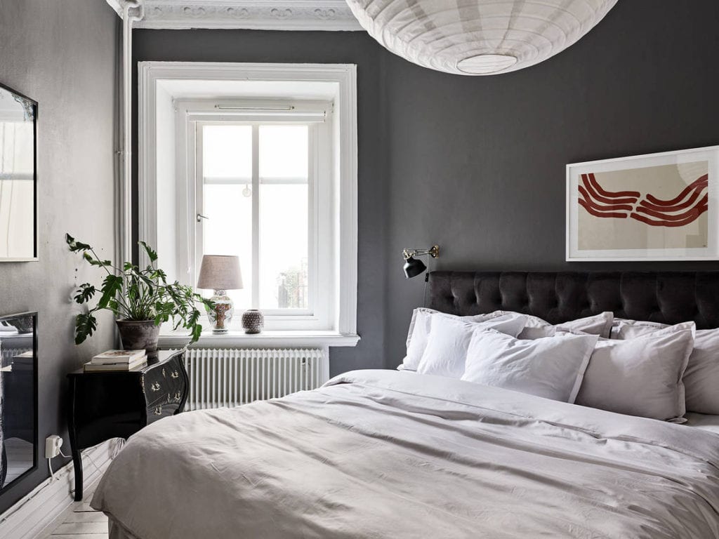 A bedroom with dark grey walls, a grey velvet headboard and white and beige bedding